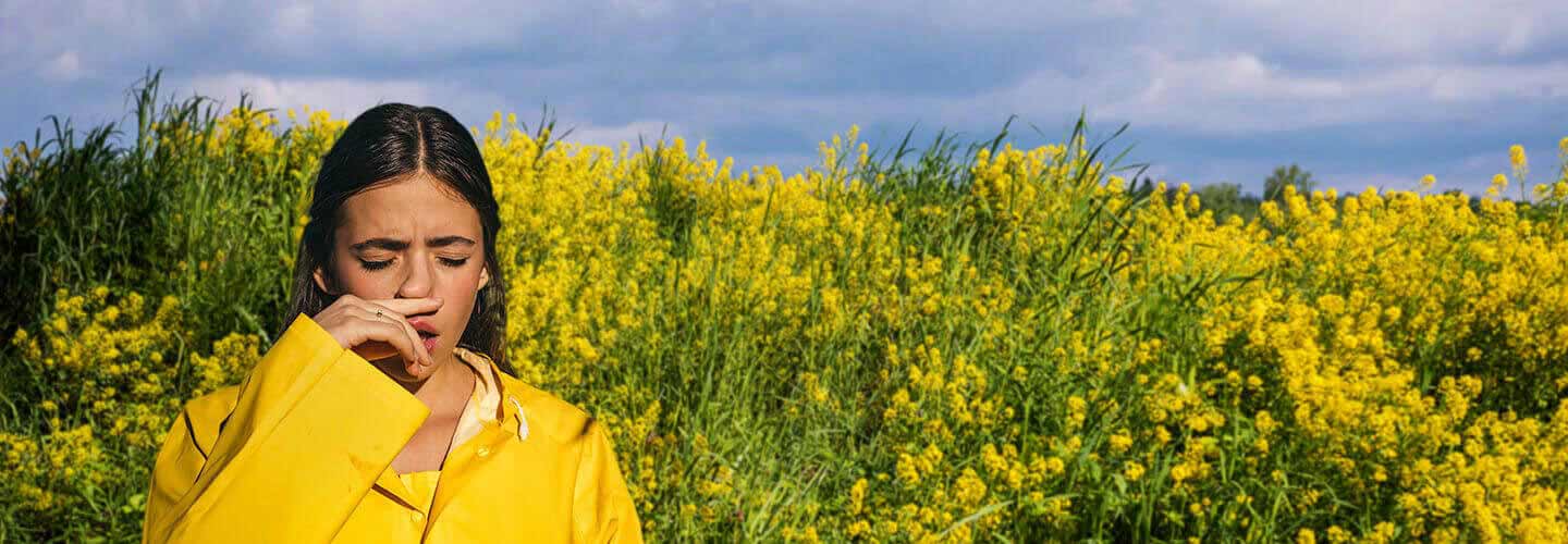 Mujer joven entre plantas floreciendo en el parque, estornuda debido a la rinitis alérgica - Rhinomer 